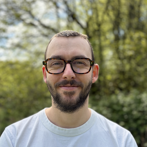 Larry standing outdoors in front of green trees. Larry is a white man in his early thirties with short brown hair, a short beard and dark-framed glasses, wearing a light t-shirt and smiling slightly.